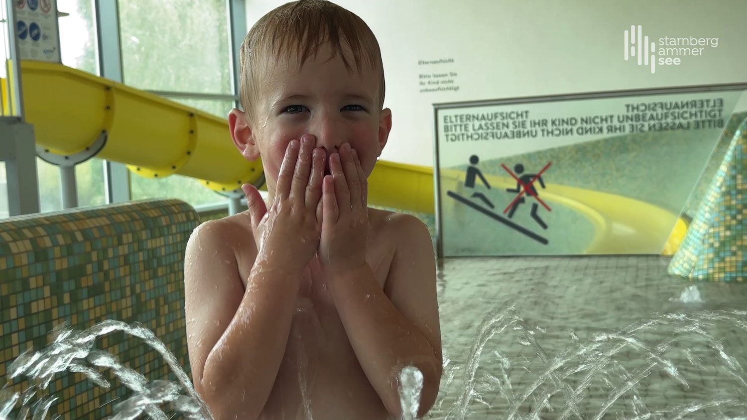 A smiling boy plays in the indoor area of a swimming pool in front of a yellow waterslide, with water fountains spraying from the floor – a joyful moment of water fun. Safety signage in the background highlights the importance of adult supervision.