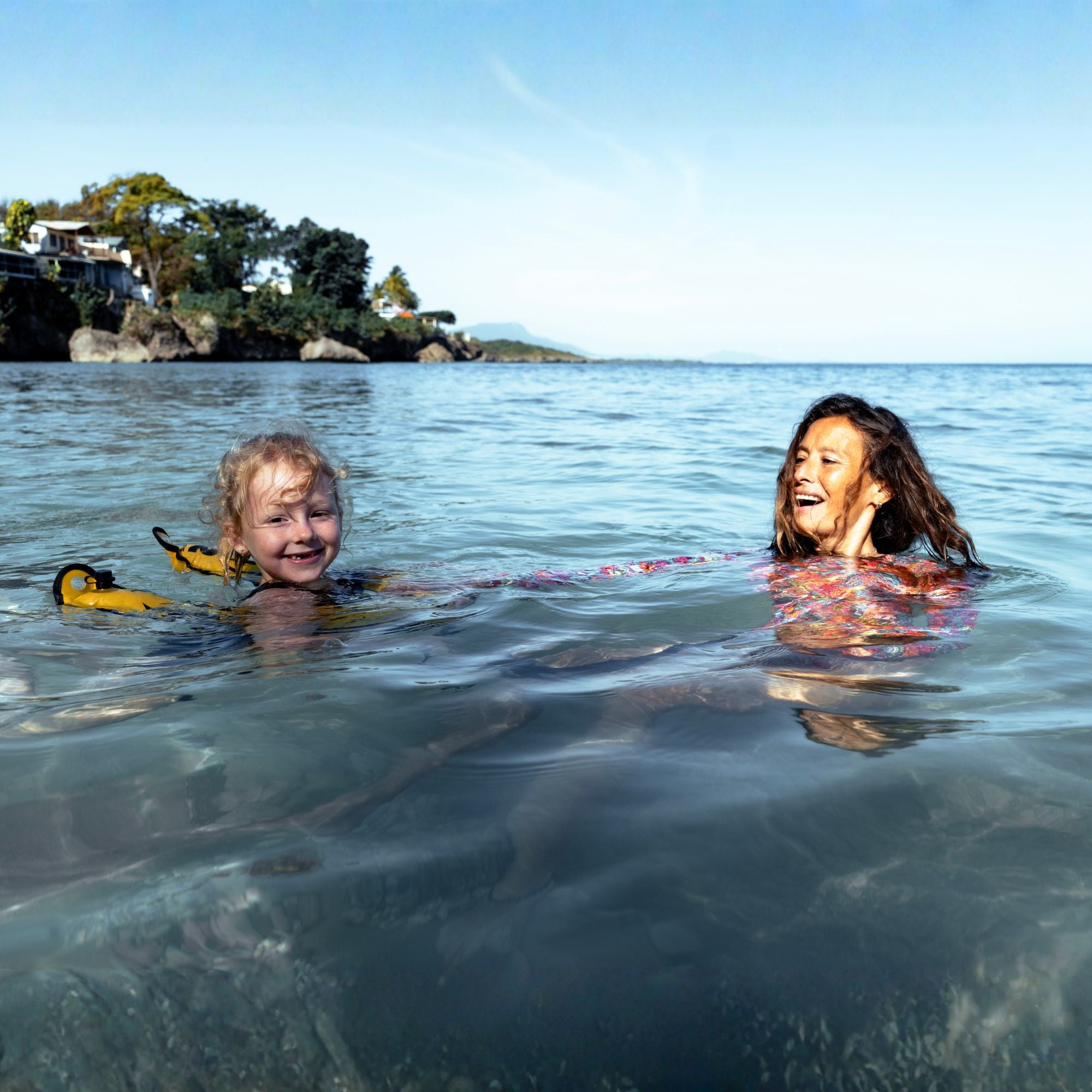 Young girl swimming in the sea with a yellow swim buoy while a woman swims beside her in shallow coastal water.
