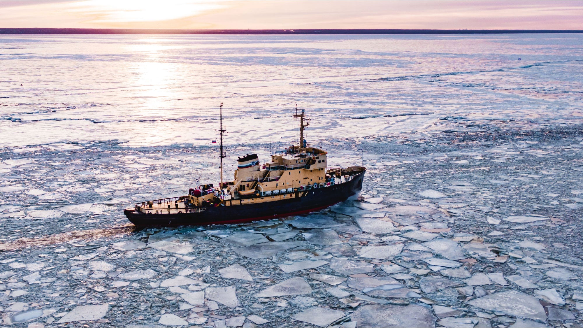 A ship navigating through broken sea ice on a frozen ocean at sunset, viewed from above.