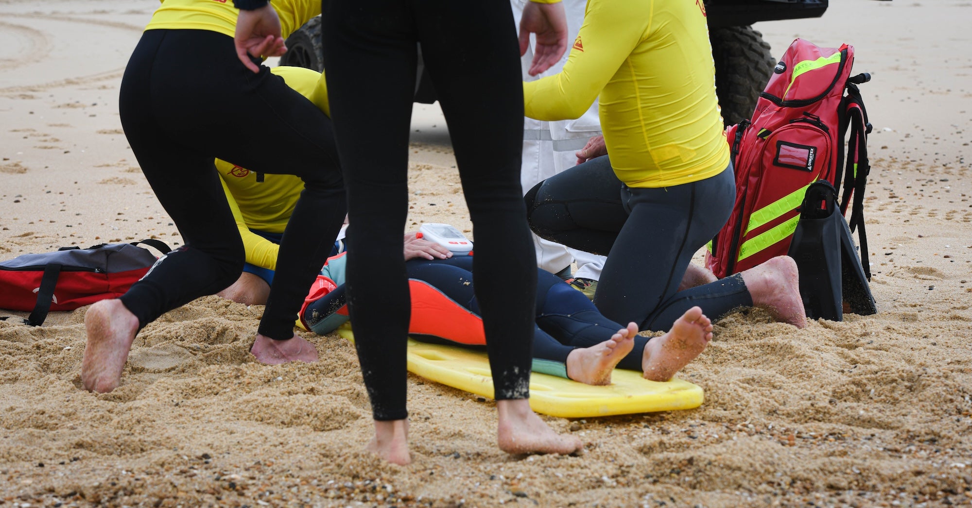 Lifeguards in yellow shirts kneeling around a person on a rescue board during a beach emergency response, with a red first aid backpack nearby