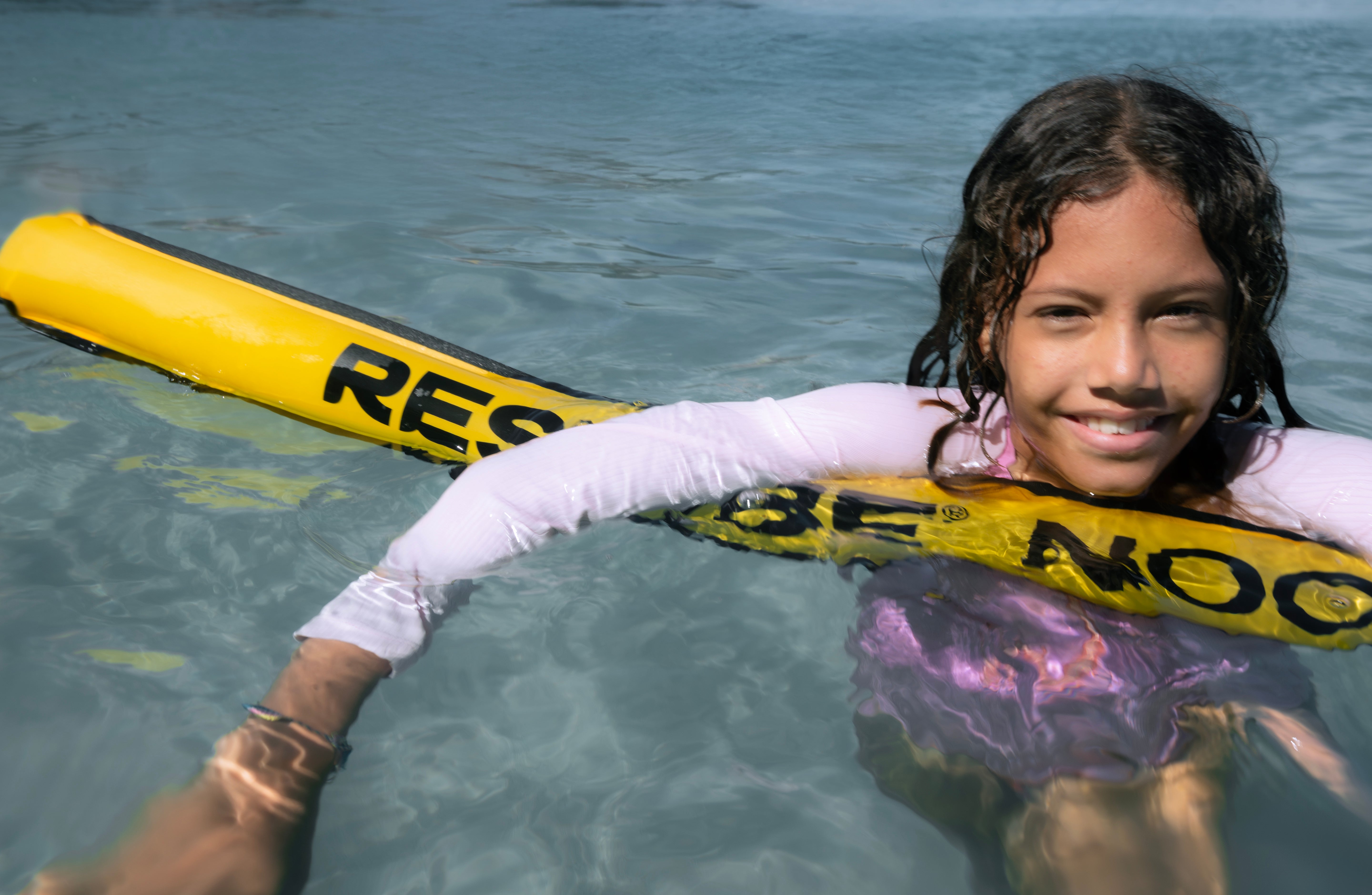 Smiling girl floating in calm blue water with a yellow pool noodle, wearing a pink and white rash guard.
