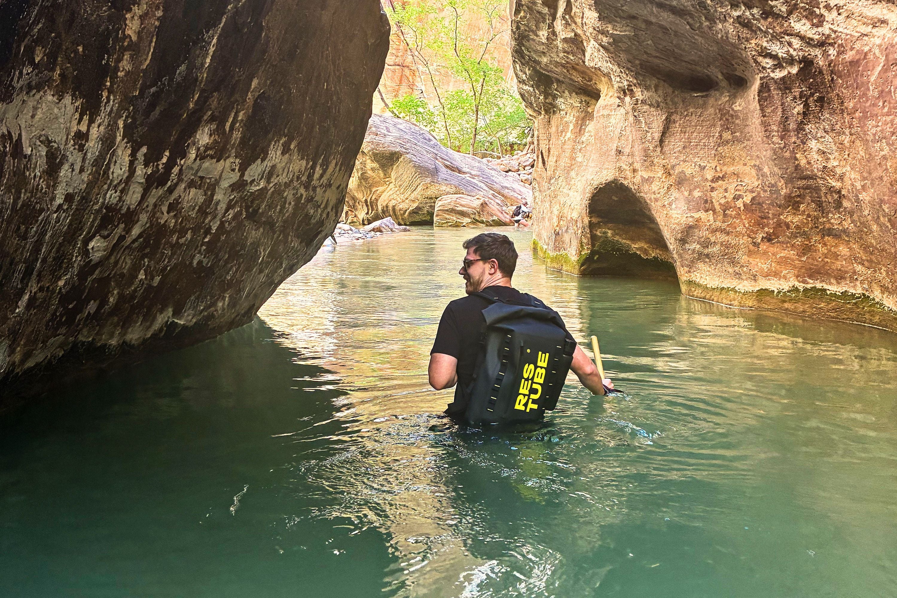 Man wading chest-deep through a slot canyon river wearing a black RESTUBE COAST water-resistant backpack, Zion Narrows