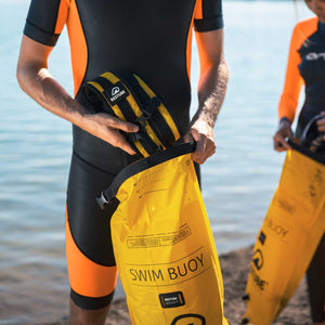 man standing at the lake holding the swimbag and putting the backpack straps into the bag