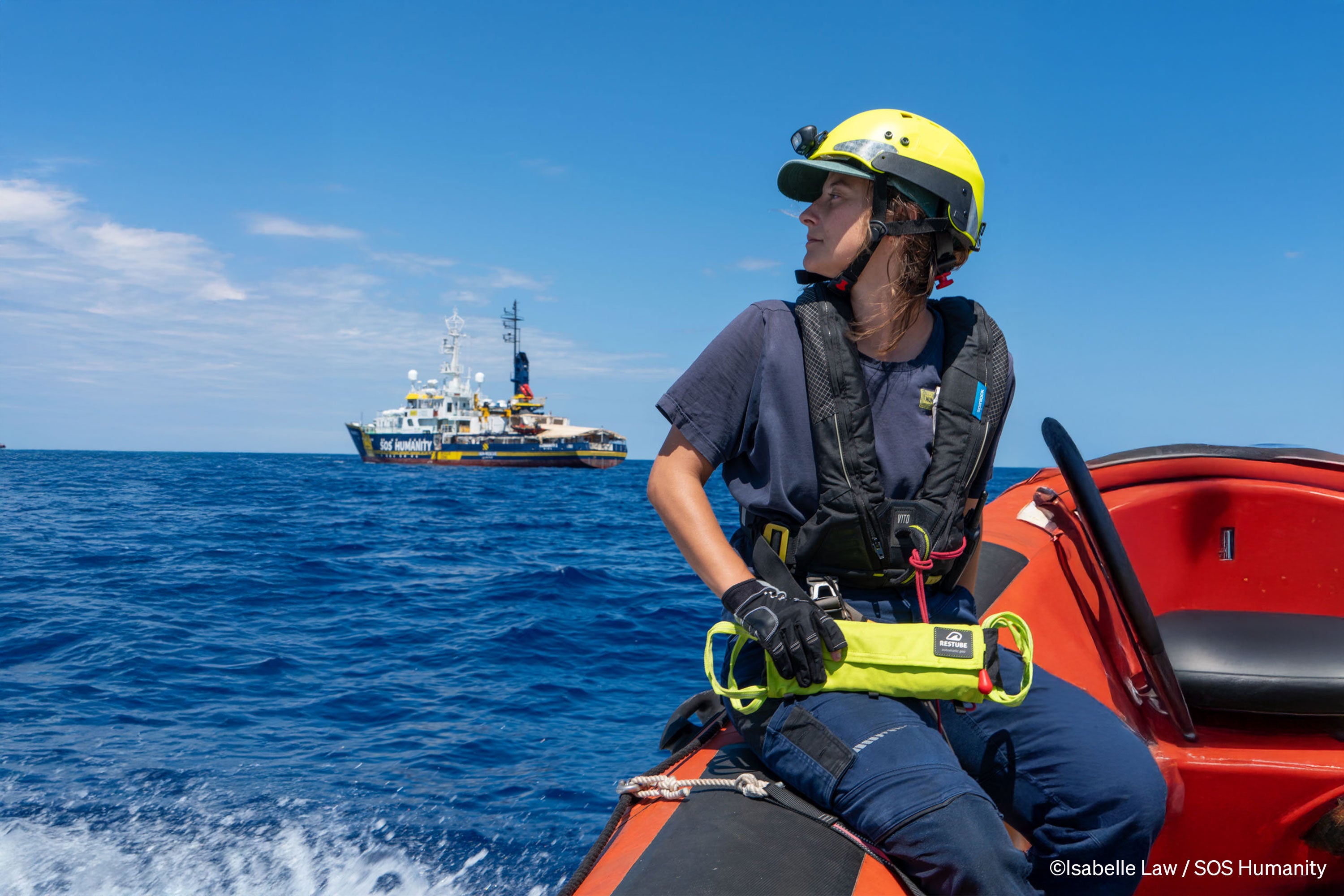 Rescue worker wearing helmet with a RESTUBE AUTOMATIC 180 sitting on an rescue boat, with the SOS Humanity rescue ship in the background on the open sea.