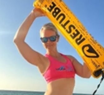 Person wearing a pink sports top and sunglasses at the beach, holding an inflated yellow RESTUBE safety buoy above their shoulder with the ocean in the background.