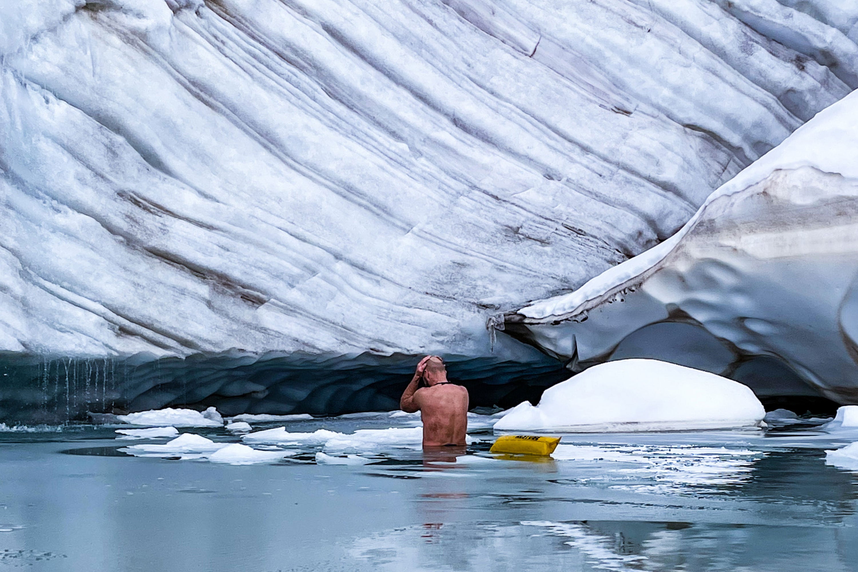 A man in icy water with yellow RESTUBE buoy in front of glacier wall.