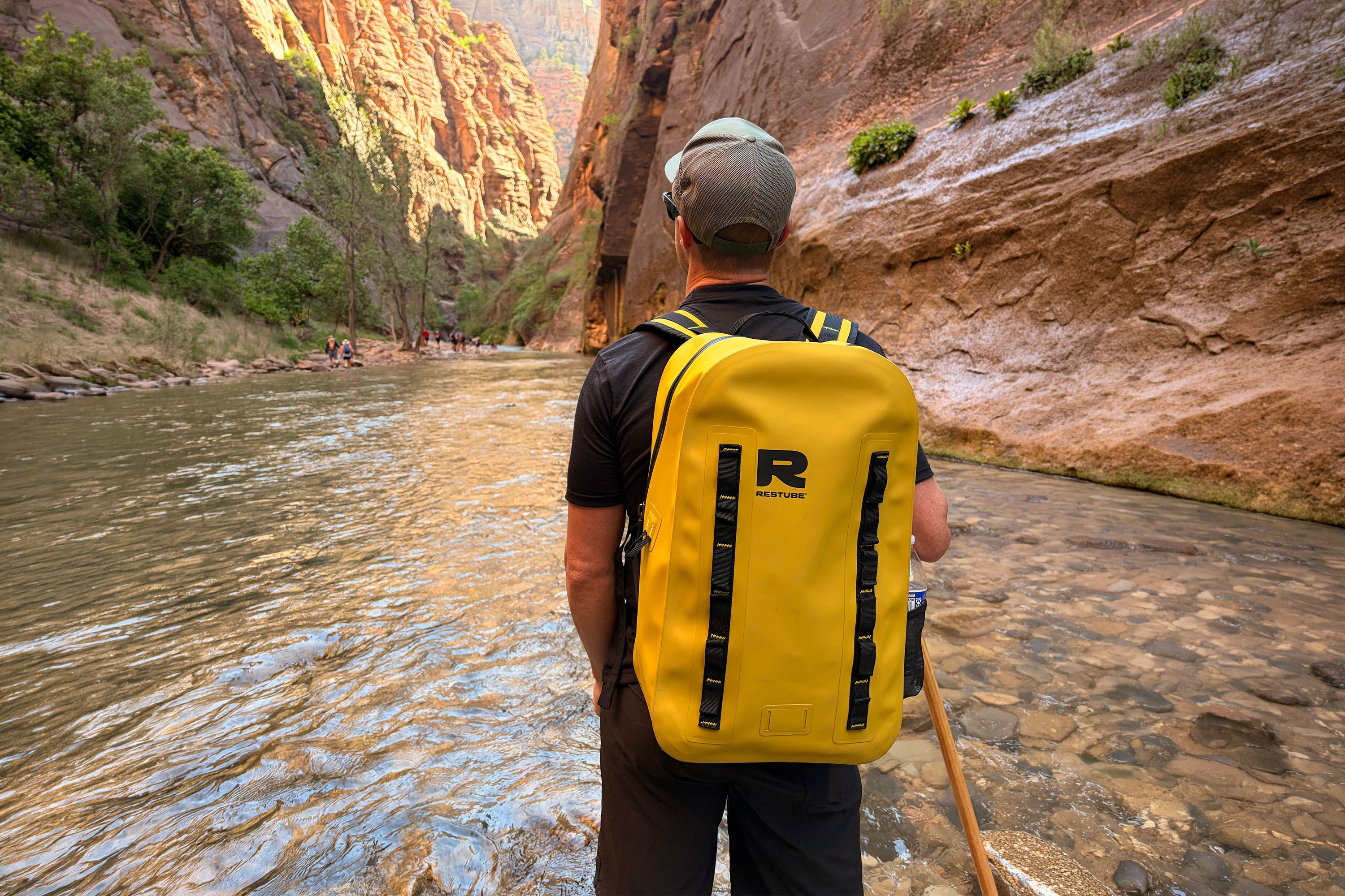 Man standing in a shallow river canyon viewed from behind, wearing a yellow RESTUBE OCEAN backpack, Zion Narrows in the background