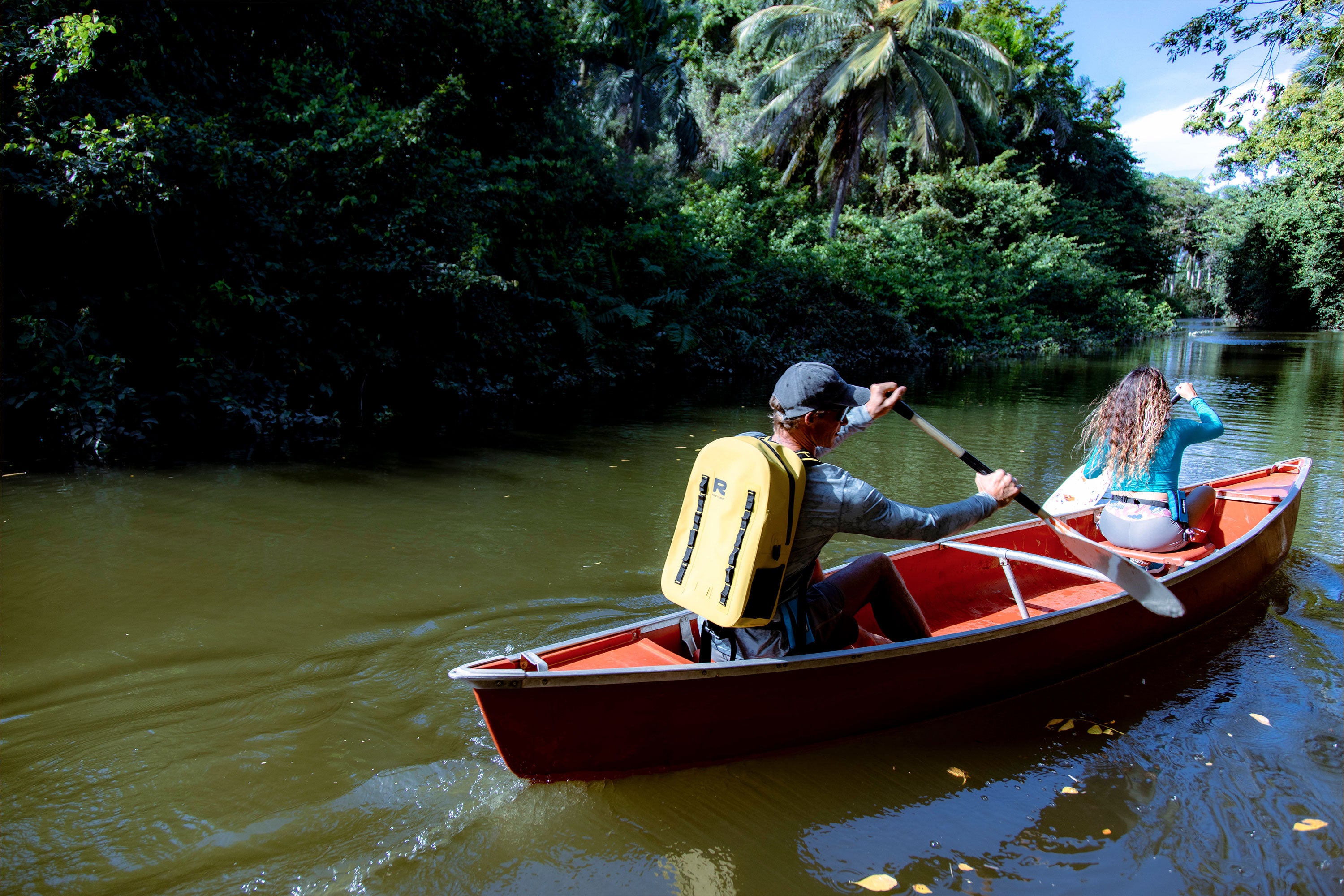 Two people paddling a red canoe through a calm jungle river, man in front wearing a yellow RESTUBE OCEAN backpack