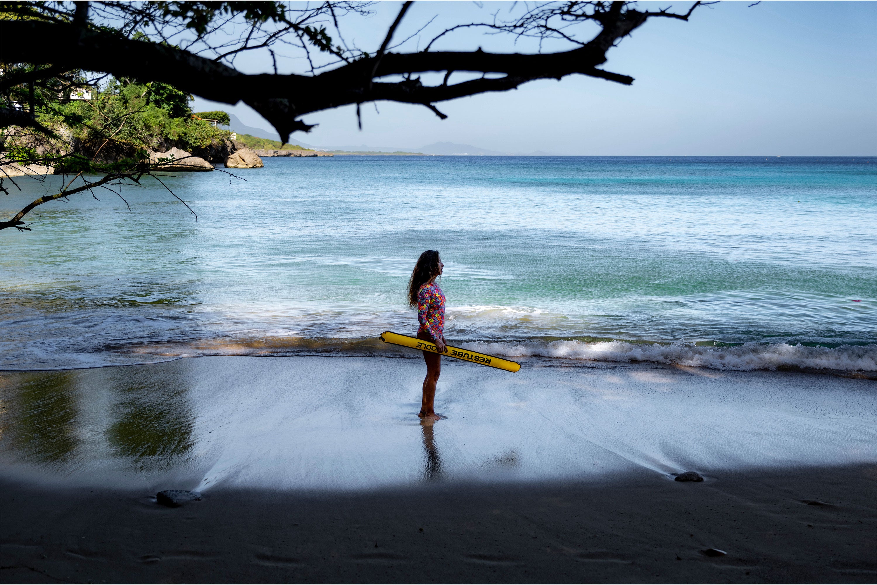 A child standing on a tropical beach holding a yellow RESTUBE Pool NOODLE near the ocean shoreline.