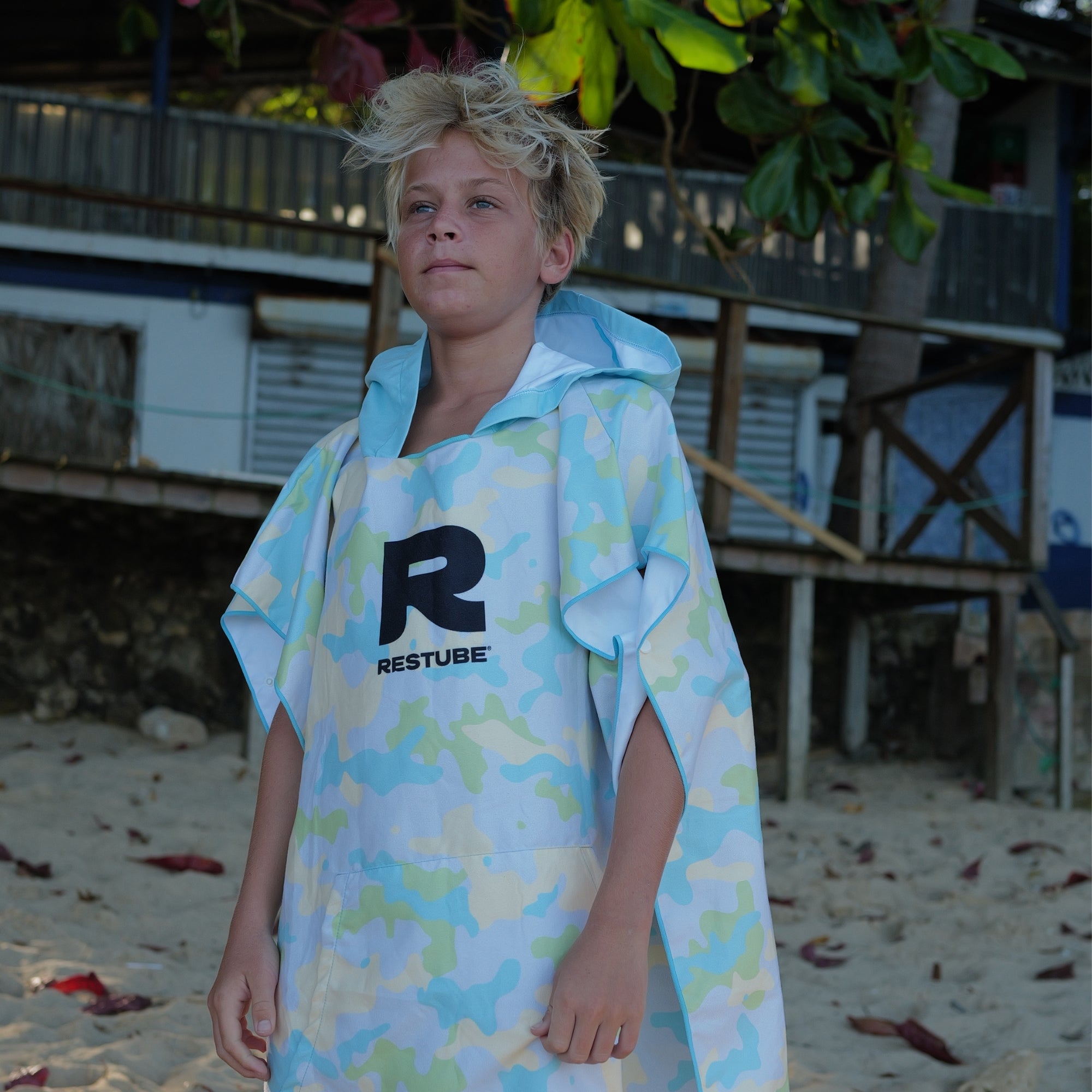 Boy wearing the RESTUBE PONCHO KIDS in light yellow camo print on a sandy beach. Hooded kids' surf poncho with RESTUBE logo on the front.
