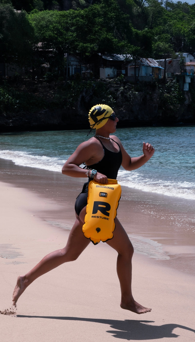 Woman in a yellow swim cap and goggles running along the beach carrying a yellow RESTUBE SWIM BUOY, with a tropical bay and hillside village in the background.