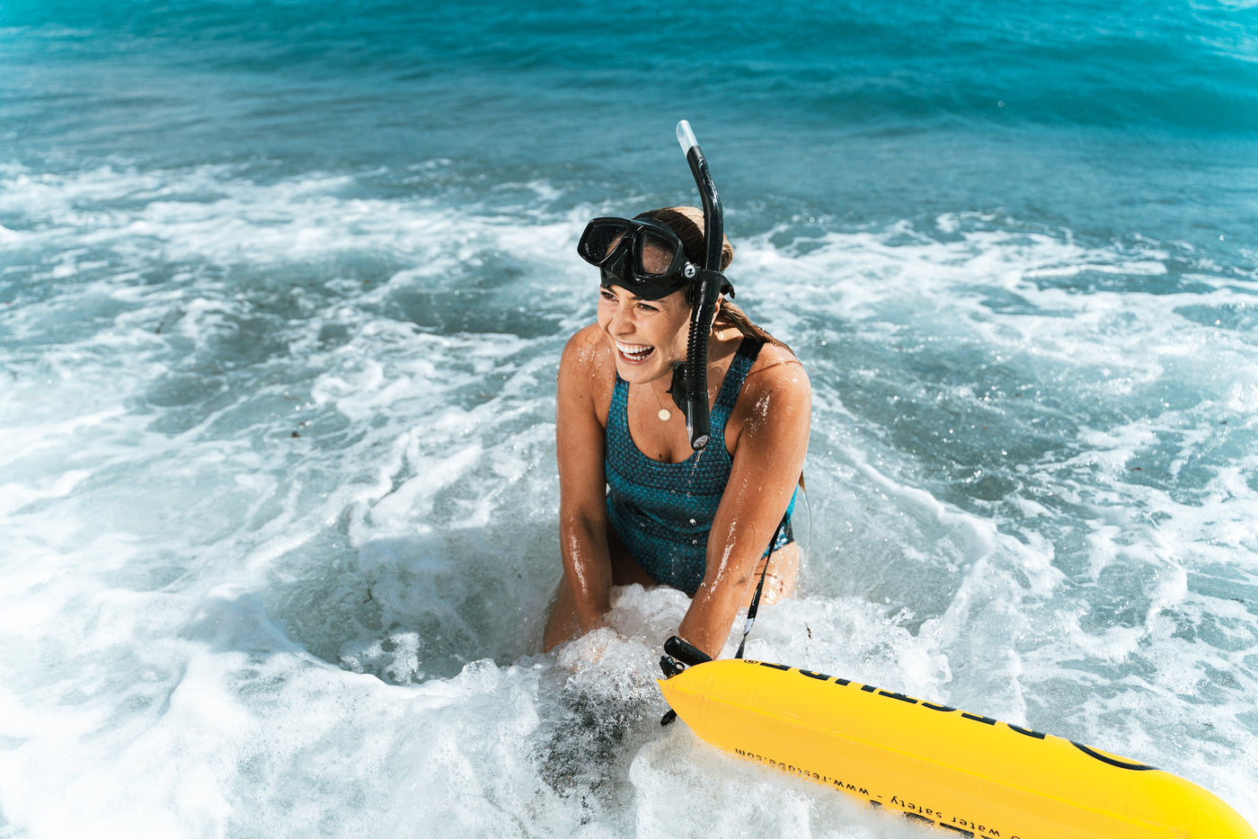 Frau mit Taucherbrille, Schnorchel und halb automatisch aufgeblasener Restube Schwimmboje kniet in den Wellen und hat Spaß.