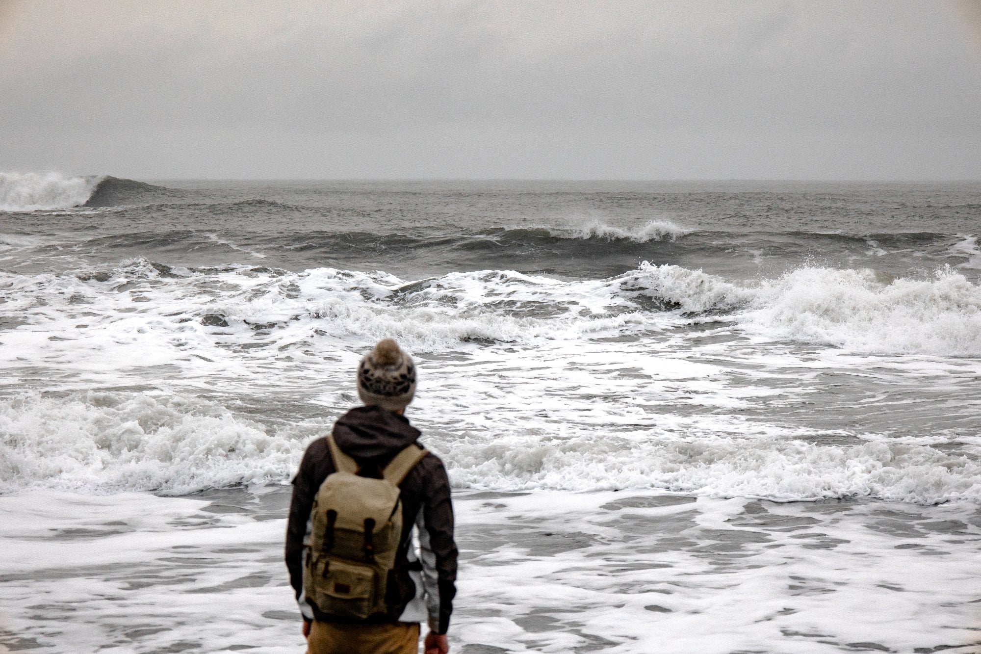 Person standing on the shore watching rough ocean waves under a grey, stormy sky.