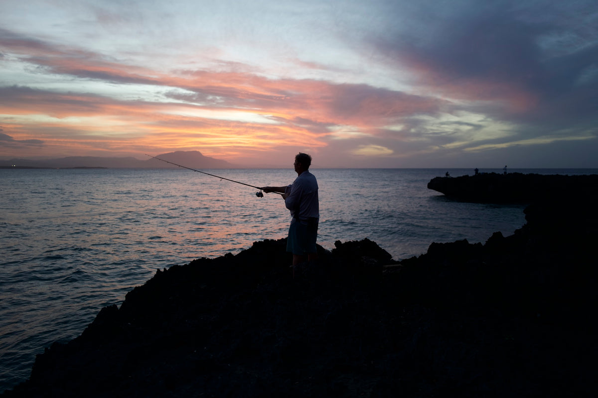Angler casting from a rocky coastline at sunset under a pink and orange sky, with other anglers on a rock ledge in the distance.