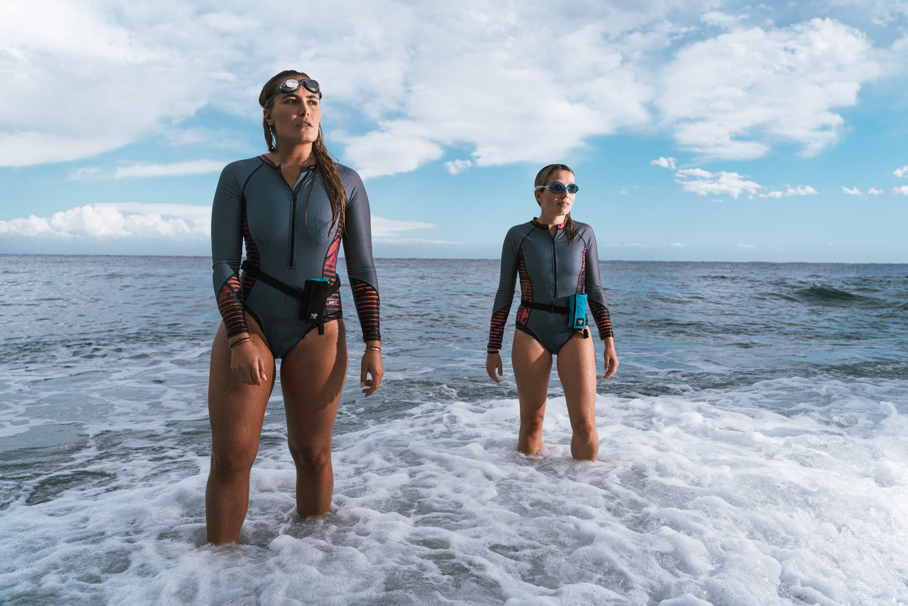 Zwei Frauen in silbergrauen Badeanzügen stehen mit Restube beach, activ und Schwimmbrillen im welligen Wasser.