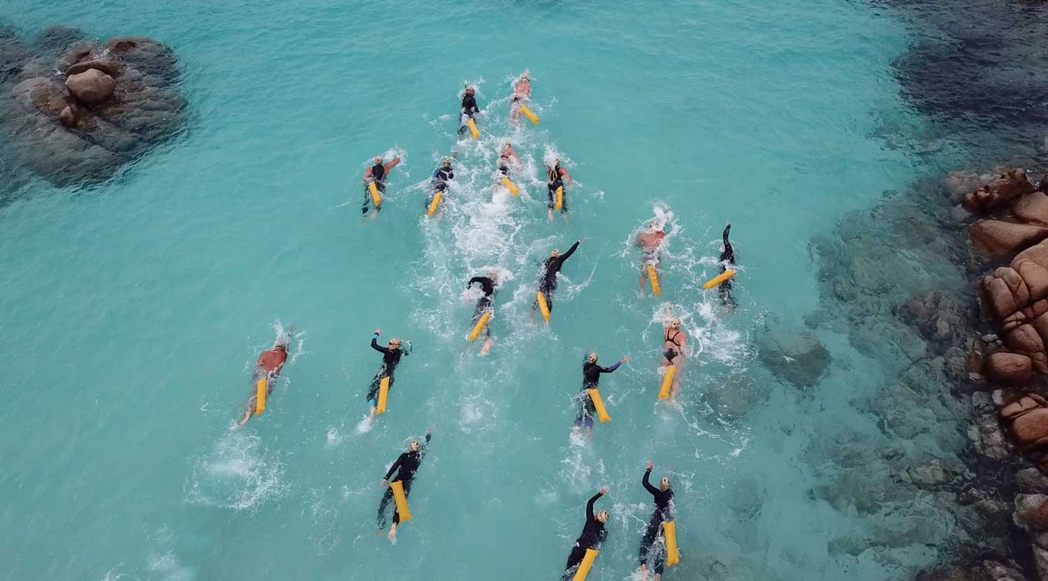 Eine Gruppe von Triathleten schwimmt mit aufgeblasenen Restube durch felsiges, hell blaues Meer.