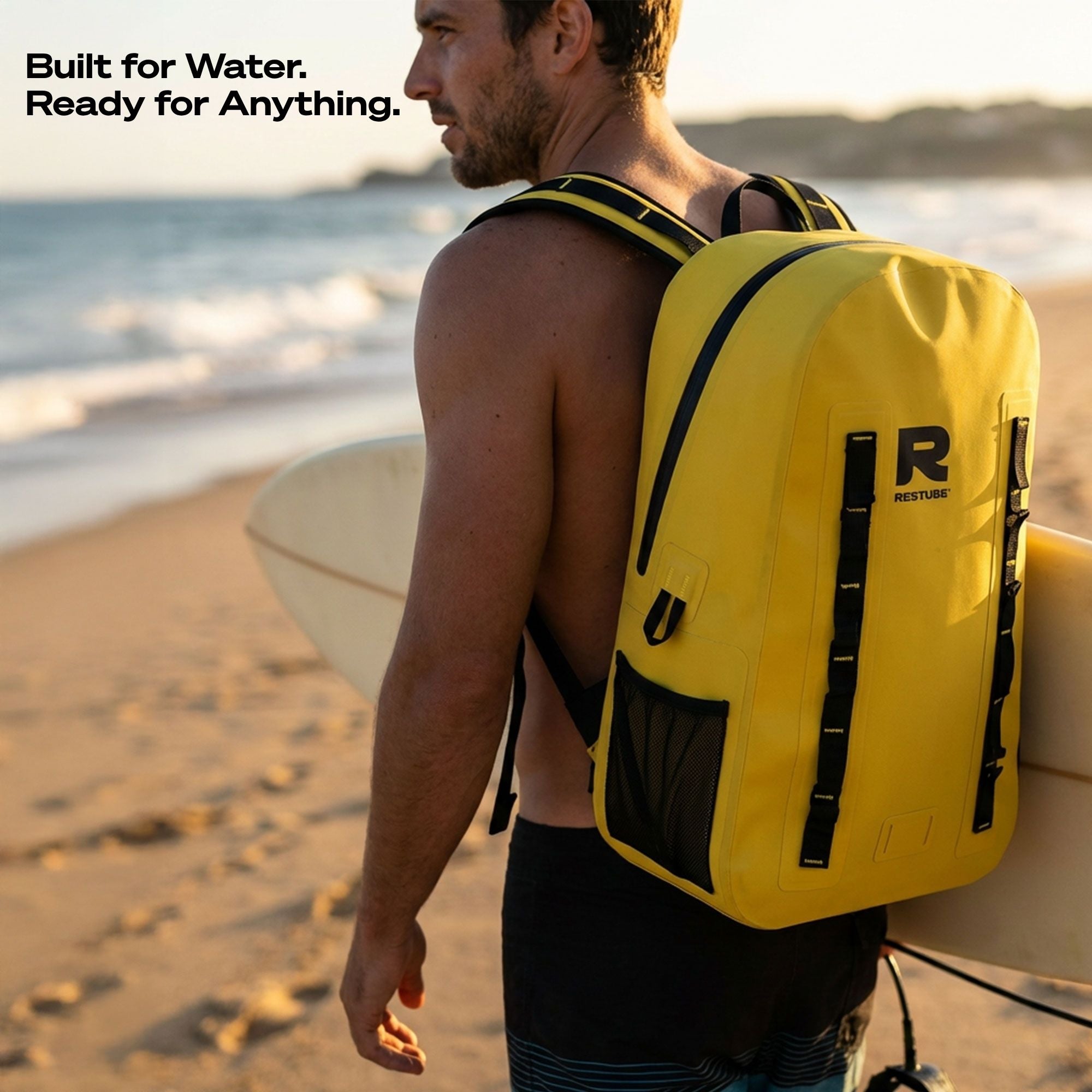 Man wearing yellow waterproof backpack on the beach holding a surfboard, showcasing outdoor use by the water