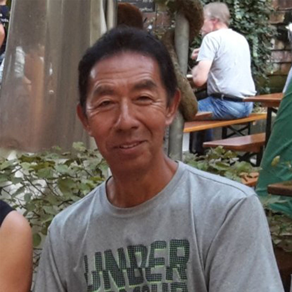 Older man sitting relaxed at an outdoor café or beer garden, wearing a gray T-shirt and smiling warmly at the camera. Other people are seated at wooden tables in the background.