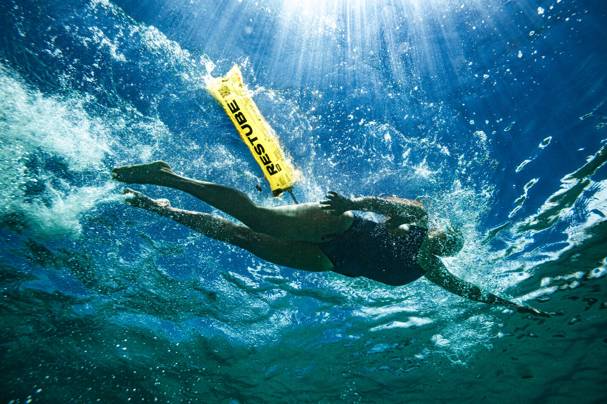 Underwater view of a swimmer diving upward toward the surface, with a yellow RESTUBE buoy above and sunlight streaming through the water