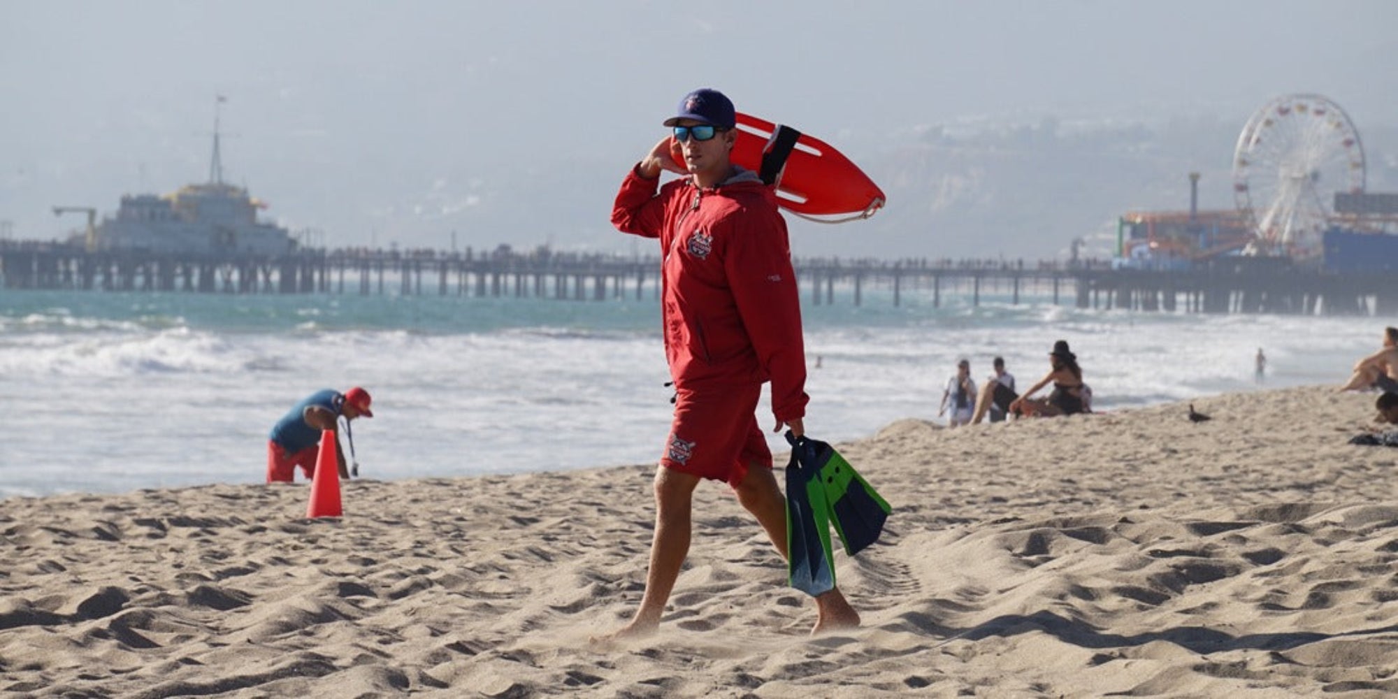Baywatch Rescue Can, Rettungsschwimmer mit Baywatch Boje und Flossen an einem Sandstrand mit Pier im Rücken.