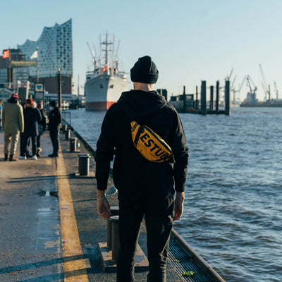 Mann am Hafen mit geschulterter Bumbag by RESTUBE X Bracenet läuft am Pier entlang
