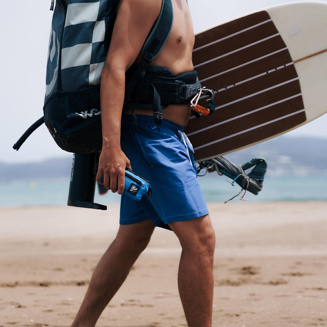 Mann mit einer Restube extreme Schwimmboje in der Hand und Kiteboard unter dem anderen Arm läuft am Strand entlang.